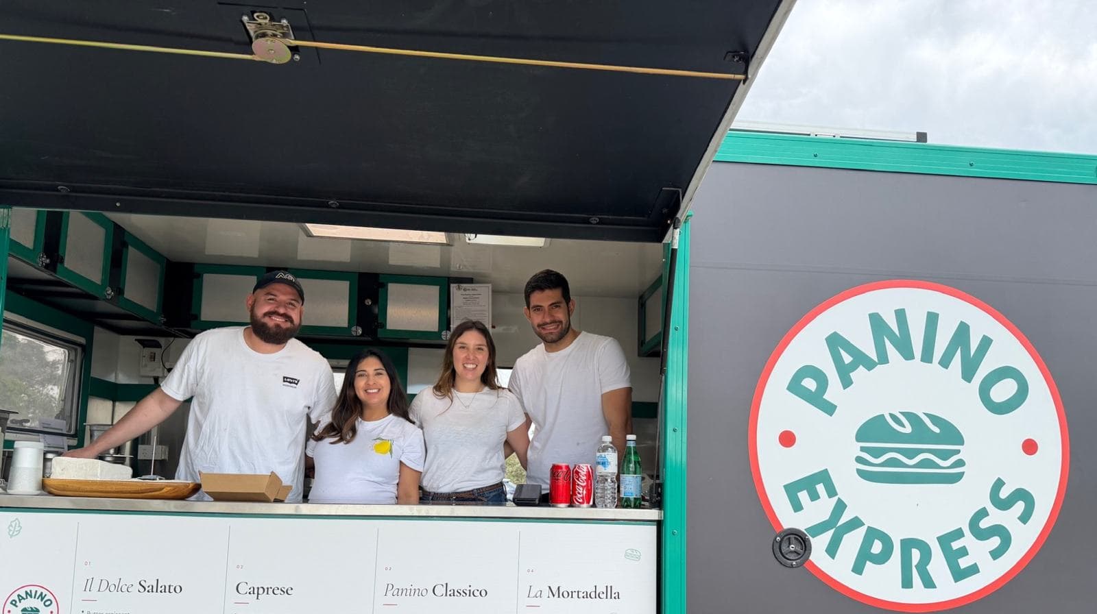 Four of the Panino Express team smiling behind the open serving hatch of the trailer, next to the roundel